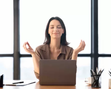 A female employee showing workplace wellness as she works in an office, meditating as her desk, feeling peaceful.