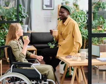 Two young intercultural employees discussing working points at lunch break while having snacks and coffee in a modern design office space surrounded by plants.